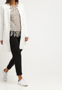 Manteau long blanc sur un pull beige texturé, pantalon noir et baskets blanches. Détail notable de franges sur l'ourlet du pull.