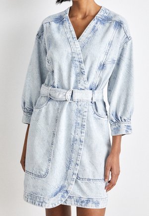 Nainen pukeutuneena vaaleansiniseen happopesuun farkkumekkoon, jossa on pitkäpuffihihat, vyö ja etutaskut.