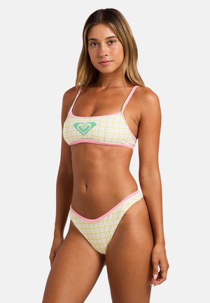 Mladá žena nosí žlto-ružové kockované bikiny s ružovým lemom a zeleným logom na vrchu, stojí pred jednoliatym svetlým pozadím.