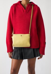 Borse a tracolla gialla in pelle con finitura testurizzata, hardware dorato e logo in rilievo, indossata sopra un maglione rosso in maglia e una gonna a quadri.