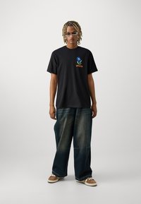 Camiseta de algodón negra con un diseño floral azul y texto naranja. Combinada con pantalones de mezclilla azules holgados y zapatos acentuados en marrón.
