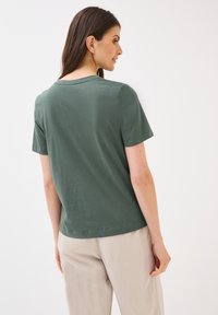 En kvinna står vänd bort, iklädd en grön t-shirt och beige byxor, som visar en avslappnad passform och enkel stil mot en enkel vit bakgrund.