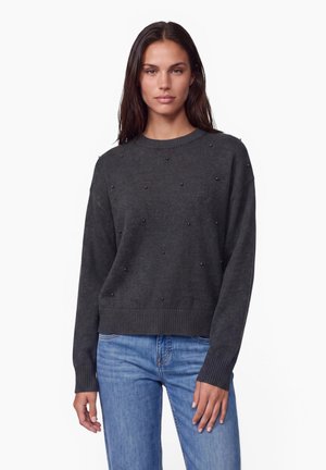 Femme portant un pull gris foncé avec de petites perles décoratives et un jean bleu clair, se tenant devant un fond blanc uni.