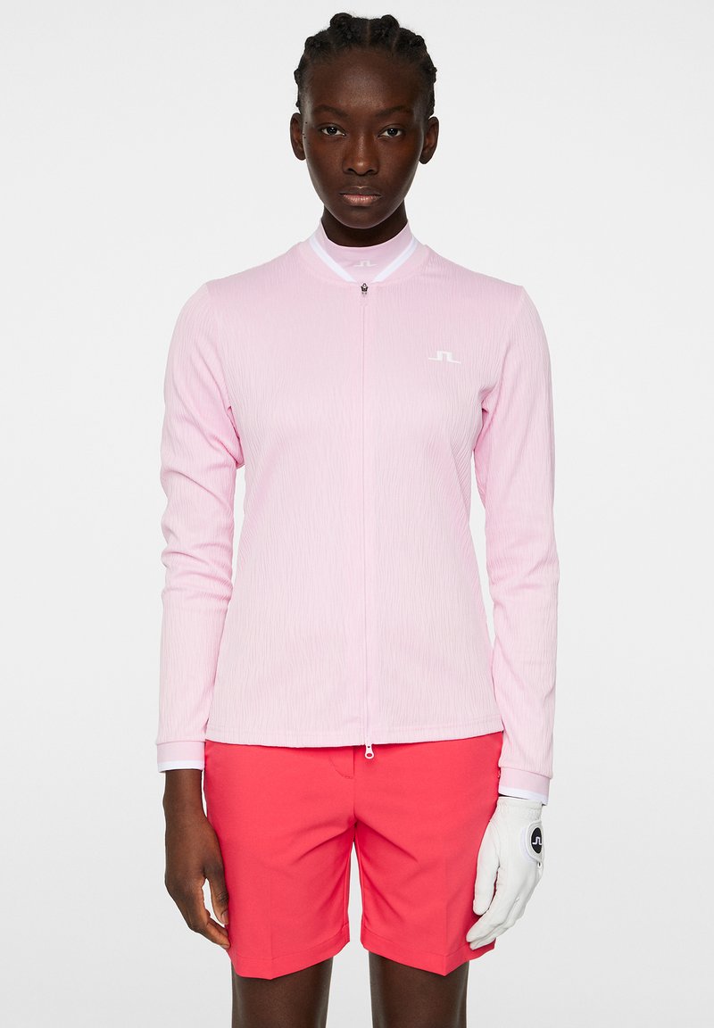 Långärmad zip-up skjorta i ljusrosa med texturerat material och vita accenter, matchad med korallfärgade shorts och en vit handske.