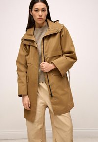 Veste imperméable marron avec capuche, fermetures à pression et fermeture éclair, trois poches avant et coupe décontractée. Portée par-dessus un pull gris et un pantalon beige.