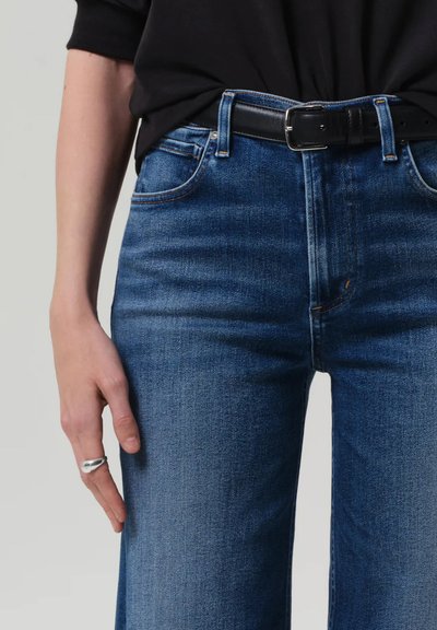 Kék farmernadrág slim fit szabással, öt zsebes dizájnnal és kontrasztos varrással. Fekete bőrövet visel. Sima textúra.