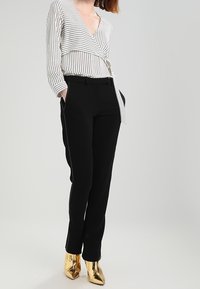 Schwarze, maßgeschneiderte Hosen mit schmaler Passform und seitlichem Streifendetail, kombiniert mit einem weiß-navy gestreiften Wickeltop. Goldene, spitze Stiefeletten.