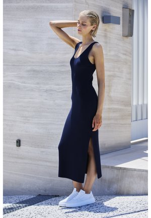 Marineblaues Strickkleid mit ärmellosem Design, hohem Schlitz und taillierter Silhouette, kombiniert mit weißen Sneakers vor einem strukturierten Hintergrund.