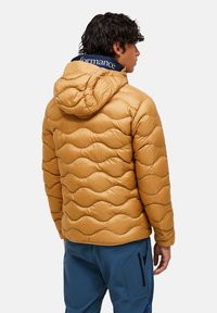 Ung mann iført en beige bølgete quilted hettejakke og blå bukser, stående med ryggen til mot en ensfarget bakgrunn.