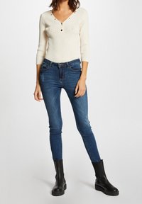 Cremefarbenes, geripptes Langarmshirt mit V-Ausschnitt und Knopfdetails, kombiniert mit dunkelblauen Skinny-Jeans und schwarzen Stiefeletten.