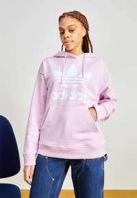 Lys rosa hettegenser med stor hvit Adidas-logo og tre-stripes design. Har frontlomme og snorhette. Laget av bomull.
