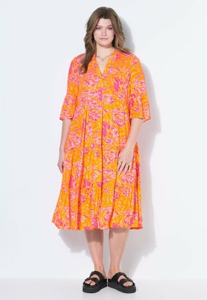 Kvinde iført en klar orange midikjole med pink blomsterprint, trekvartærmer og sorte platformssandaler, stående mod en hvid væg.