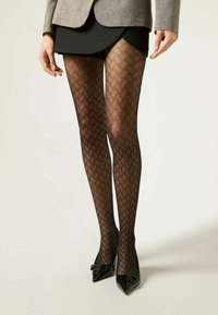 Collants noirs à motif avec un design en losanges, associés à une jupe noire et des escarpins noirs à bout pointu brillants. Veste grise en haut.