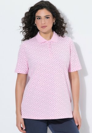 Kobieta o kręconych włosach, nosząca jasnoróżową koszulkę polo z krótkim rękawem i małymi, wielokolorowymi groszkami, stojąca na białym tle.