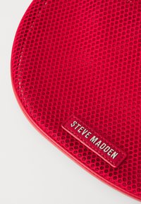 Sarkans tīkls ar sešstūra rakstiem, ar strukturētu virsmu un taisnstūra etiķeti, uz kuras baltā burtā ir rakstīts "STEVE MADDEN".