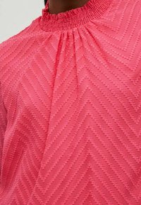 Pink blouse with a high smocked neckline, featuring a textured zigzag pattern and soft fabric.