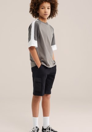 Dospievajúci v sivobielom tričku s krátkym rukávom, čiernych cargo kraťasoch, bielych ponožkách a čiernych teniskách, stojaci s rukami v náprsnom vrecku.