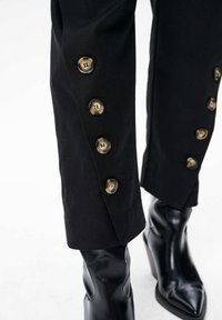 Zwarte op maat gemaakte broek met gouden knopen langs de zijkant, voorzien van een soepele stoftextuur en een slim-fit ontwerp. Zwarte haklaarzen zijn zichtbaar.