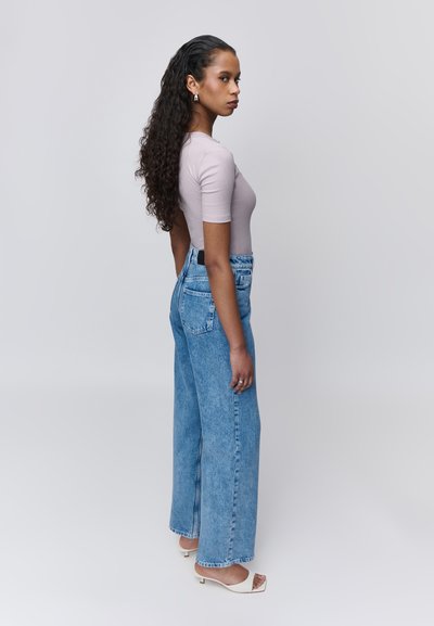 Femme aux longs cheveux bouclés portant un top moulant mauve clair, un jean bleu taille haute à jambes larges, et des sandales blanches à talons, debout de profil.