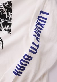 Λευκό πουκάμισο μακρυμάνικο με μαύρο γραφικό στο μπροστινό μέρος και μοβ κείμενο κατά μήκος του μανικιού που γράφει «LUXURY TO BURN». Μαλακό βαμβακερό ύφασμα.