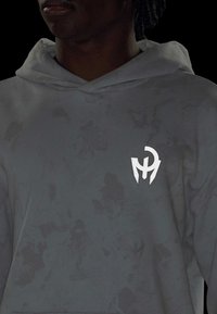 Šedá mikina s kapucňou a textúrovaným vzorom, s bielym logom na ľavom prsníku. Tkanina vyzerá mäkká s uvoľneným strihom a šnúrkami.