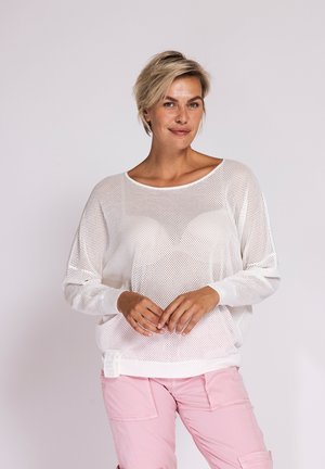 Woman with short blonde hair wearing a loose white mesh sweater and light pink cargo pants, standing against a plain background.