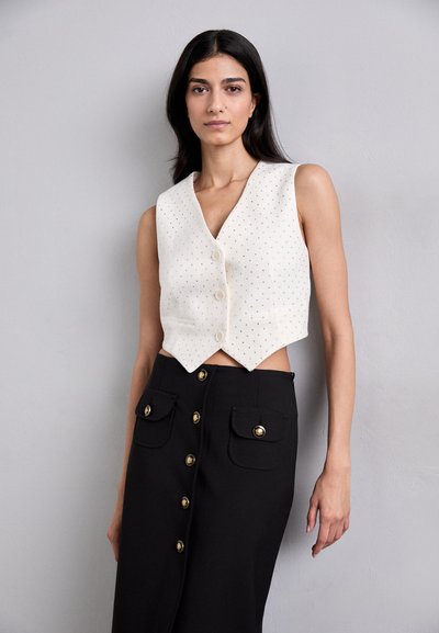 Weißes, kürzer geschnittenes Top mit V-Ausschnitt, gepunkteter Textur und vorderem Knopfverschluss, kombiniert mit einem schwarzen Rock mit goldenen Knopfdetails.