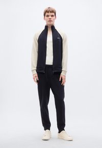 Μπλε ναυτικό μπουφάν με φερμουάρ και μπεζ μανίκια, σετ με τζόκερ παντελόνια στον ίδιο τόνο. Διαθέτει μαλακό ύφασμα, πλαϊνές ρίγες και κρεμ αθλητικά παπούτσια. Απλός σχεδιασμός.