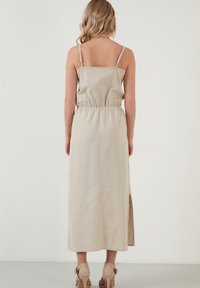 Béžové maxi šaty z lněné tkaniny s nastavitelnými ramínky, vypasovaným pasem a bočním rozparkem, které mají jednoduchý design a přirozenou texturu.