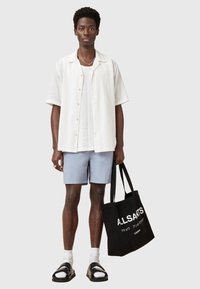 Maglietta bianca a maniche corte con una leggera texture, pantaloni corti azzurri, sandali neri e una borsa nera con testo bianco.
