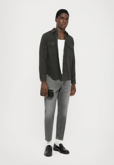 Young man standing wearing a black denim shirt over white tank top, gray jeans, white socks, black loafers, holding a black wallet in hand.