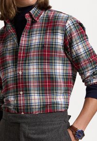 Camicia a quadri rossa, verde, bianca e blu con colletto e polsini con bottoni. Indossata con pantaloni grigi e un orologio da polso in pelle marrone.