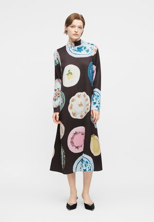 Moteris stovi vilkėdama ilgomis rankovėmis juodą suknelę su ryškiais dideliais gėlių ir plokščiomis apskritomis kaitomis, derinama su juodais smailiais bateliais.