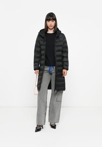 Lang zwarte gewatteerde jas met een capuchon, geborduurd ontwerp, gecombineerd met grijze wijde jeans en zwarte puntige hakken. Lichtroze handtasje als accessoire.