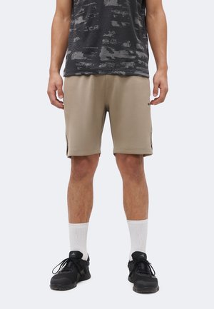 Mand iført beige knælange shorts, hvide ribstrikkede crew-sokker, sorte sneakers og en sort mønstret kortærmet skjorte, stående foran en ensfarvet baggrund.