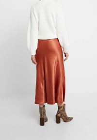 Jupe midi en satin rouille avec une texture lisse, associée à un pull en maille épaisse blanc et des bottines à talon bloc en peau de serpent marron.