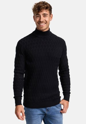 Zwarte gebreide trui met een hoge kraag, met een golfpatroon en geribde manchetten. Gestyled met blauwe spijkerbroeken.