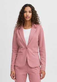 Růžový blazer se strukturovaným designem, zapínáním na jeden knoflík a dvěma předními kapsami. Hladká textura a přiléhavý střih. Nosený přes bílý top.