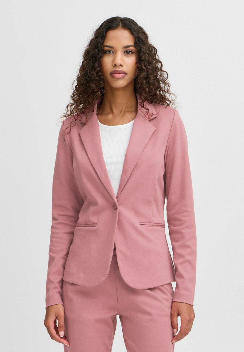 Růžový blazer se strukturovaným designem, zapínáním na jeden knoflík a dvěma předními kapsami. Hladká textura a přiléhavý střih. Nosený přes bílý top.
