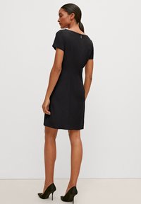 Kurzes schwarzes Kleid mit kurzen Ärmeln, taillierter Passform und einem Reißverschluss im Rücken. Glattes Material und knielanger Saum. Getragen mit schwarzen High Heels.