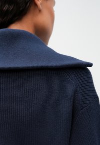 Tmavomodrý pletený sveter s širokým, prehnutým golierom, s ryhovanou textúrou a tesne priliehavým strihom.