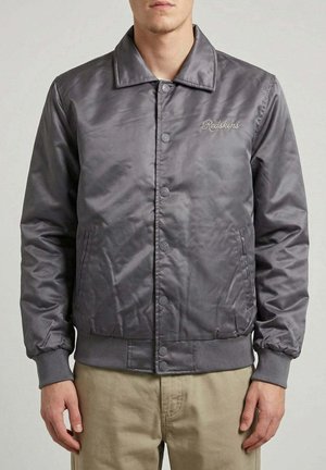 Homme portant une veste en satin grise boutonnée avec "Redskins" brodé sur la poitrine et un pantalon beige, debout devant un fond uni.