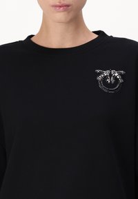 Melnā džemperis ar teksturētu, rotātu brošu ar sudraba akcentiem un pērlītēm kreisajā krūšu daļā. Klasiska apaļa kakla izgriezuma dizains.