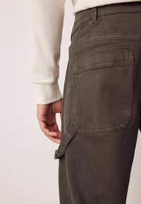 Olivgrüne Cargo-Hosen mit Seitentasche, schmaler Schnitt und subtiler Textur. Kombiniert mit einem hellen Langarmshirt.