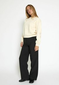 Donna che indossa una felpa color crema e pantaloni larghi neri, in piedi con le mani rilassate, guardando leggermente verso l'alto contro uno sfondo bianco.