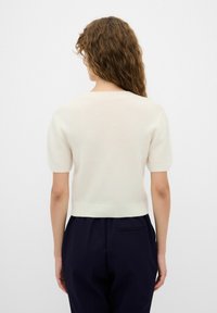 Strickpullover in Off-White mit kurzen Ärmeln, rundem Ausschnitt und tailliertem Saum. Glatte Textur; über einer dunklen Hose getragen, von hinten gezeigt.