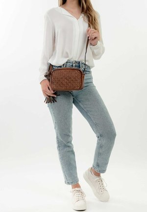 Brown textured leather crossbody bag with a geometric pattern and tassel accent, worn with light wash jeans and a white blouse.