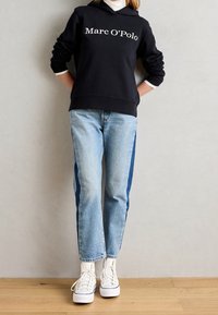 Czarna bluza z kapturem z białym logo "Marc O'Polo", zestawiona z jasnoniebieskimi dżinsami z ciemnoniebieskimi bocznymi panelami i białymi sneakersami.