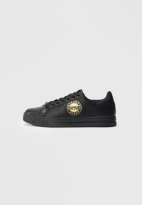 Črne nizke teniske z zlatim logotipom "Versace Jeans Couture" na strani in vtisnjeno blagovno znamko na podplatu, prikazane v profilu na beli podlagi.