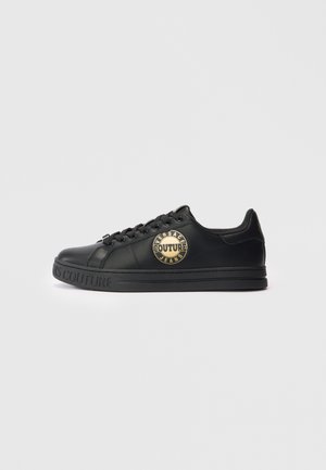 Schwarze Low-Top-Sneaker mit goldfarbenem "Versace Jeans Couture"-Logo an der Seite und geprägtem Markenlogo auf der Sohle, im Profil vor weißem Hintergrund dargestellt.